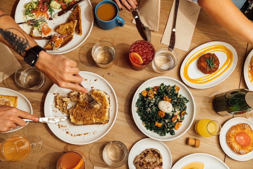 A table filled with different dishes from Paperboy with hands reaching across the table grabbing bites of food with a fork.