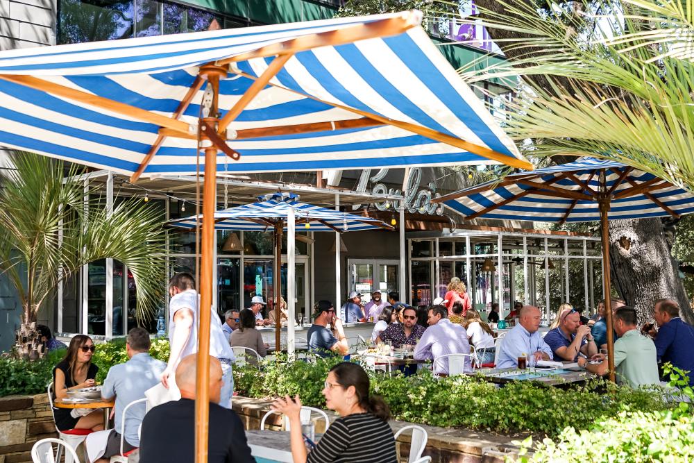 The patio at Perla's, with people dining underneath blue and white striped umbrellas and palm trees