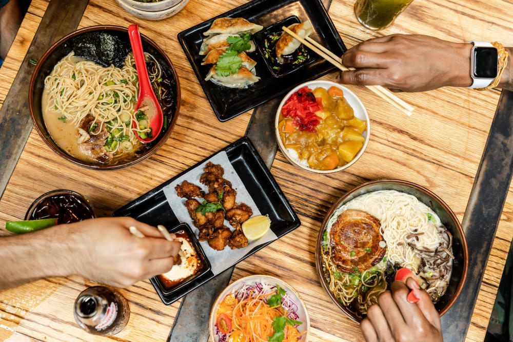 Hands reaching for different Ramen Tatsu-ya food items like ramen and crispy brussels sprouts.