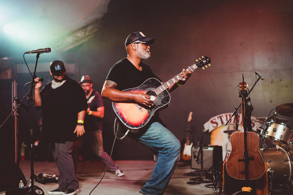 Musician Ray Prim plays an acoustic sunburst guitar on stage at Stubb's in Austin Texas. Two other male musicians stand behind him