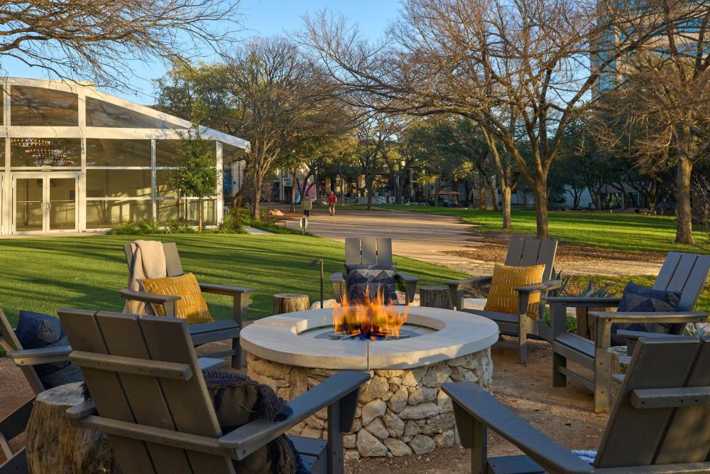 Lounge chairs around a flaming fire pit, with people walking along a path on a hotel's spacious outdoor grounds.