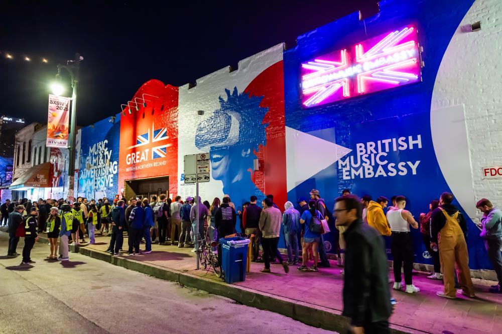 A building with large murals on the front of it with text reading "British Music Embassy" and people standing out front on the sidewalk waiting to get in.