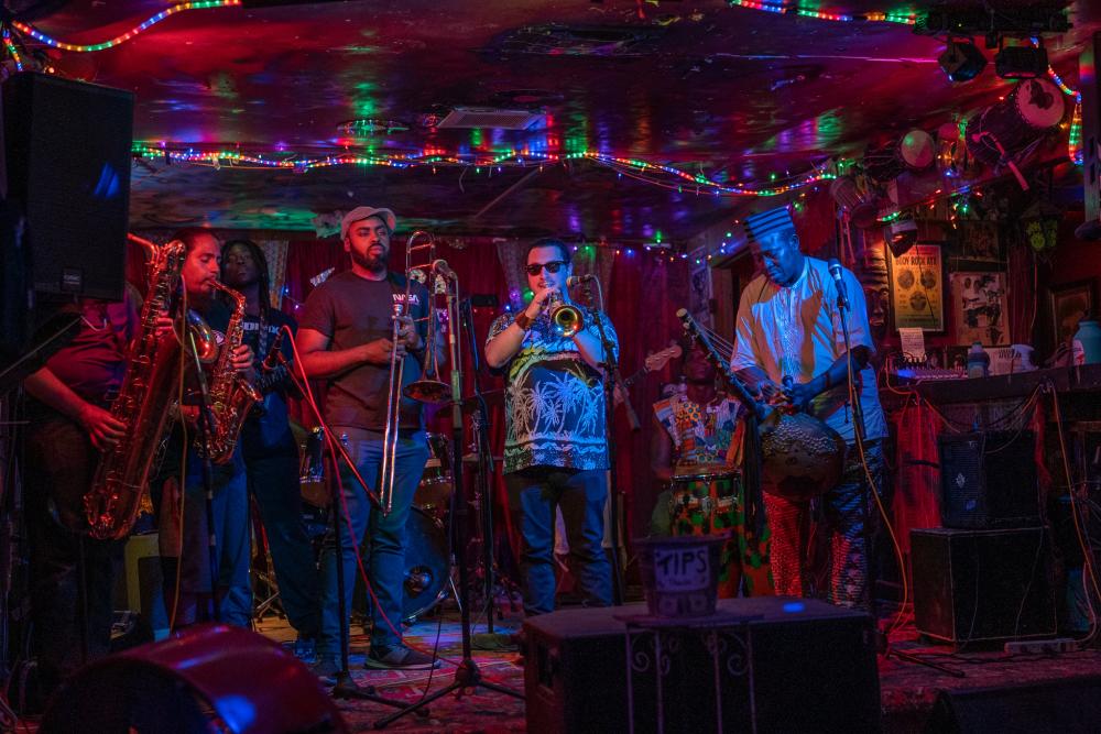 Photo of 5 men in a band on stage at the Sahara Lounge. The room is dark and rainbow rope lights surround the ceiling. From left to right person playing baritone sax, man playing alto sax, man holding trombone, man playing trumpet, man playing kora