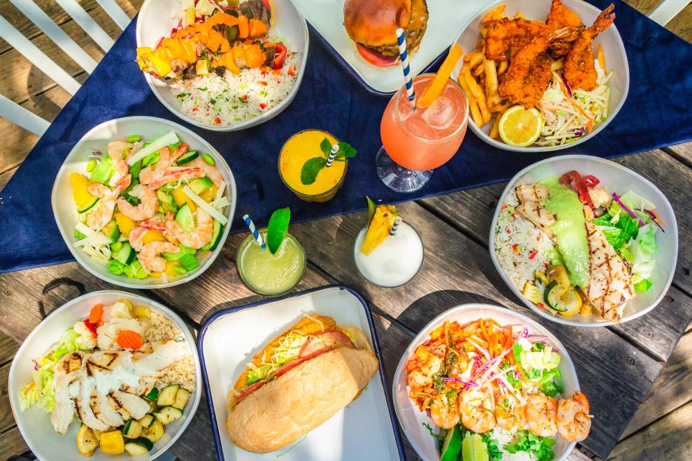 Patio table spread at Keepers Coastal Kitchen with colorful seafood dishes, a burger and three frozen cocktails