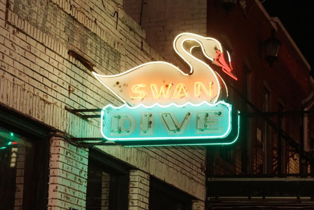 Exterior of a brick bar at night with a neon sign of a swan reading Swan Dive in white and blue lights.