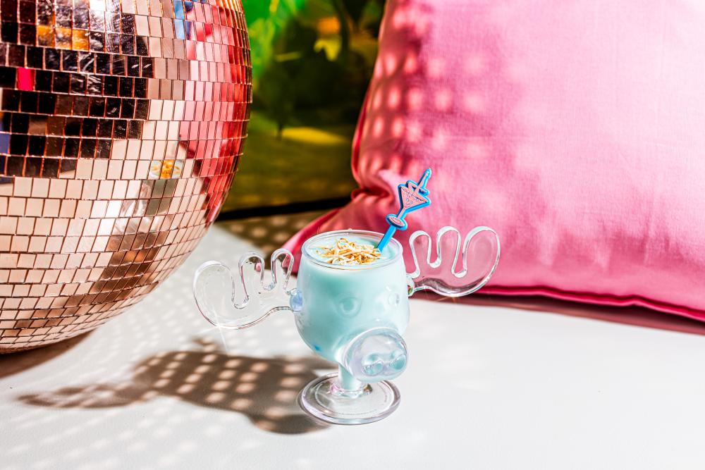 Creamy blue cocktail in a cartoon-reindeer shaped glass, sitting amongst hot pink pillows and a disco ball.