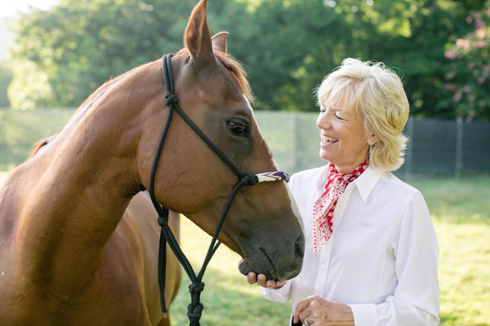 Photo of a blonde woman in a white shirt looking at a brown horse with a loose, black horse lead rope around its face