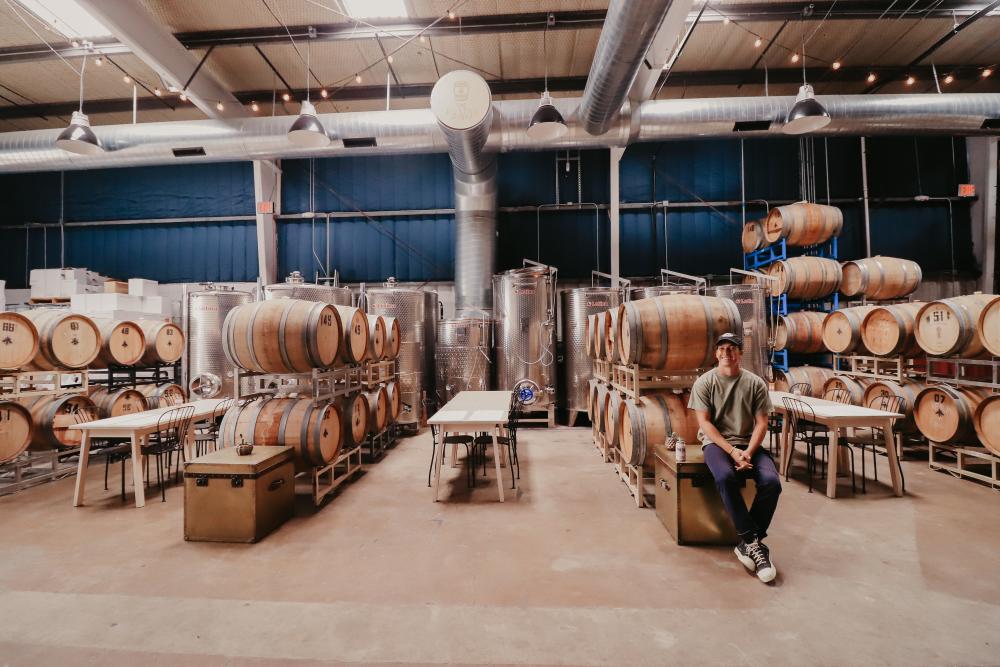 Image of the warehouse at Austin Winery with rows of barrels and the owner standing nearby smiling at the camera.