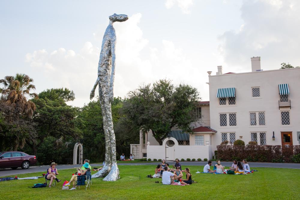 Families sitting on picnic blankets on the lawn of Laguna Gloria, circled around a large statue.