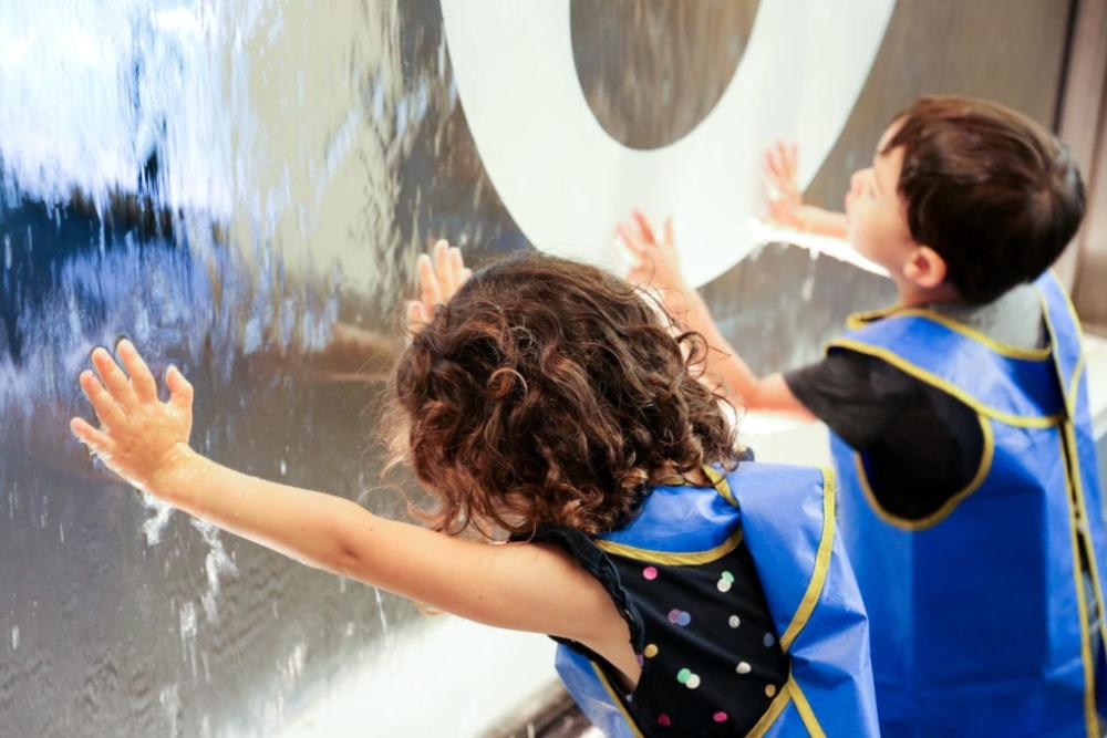 Two children in aprons pressing their hands on a glass pane with water falling down.