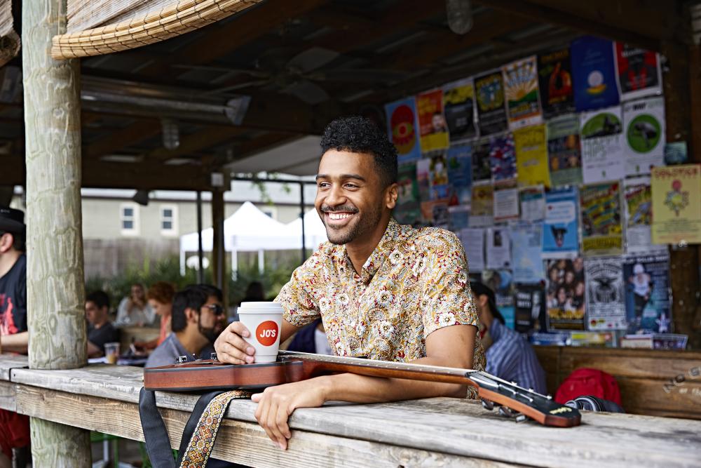 Musician Tje Austin sits on patio at Jo's South Congress with a coffee in his hand and his guitar on the table