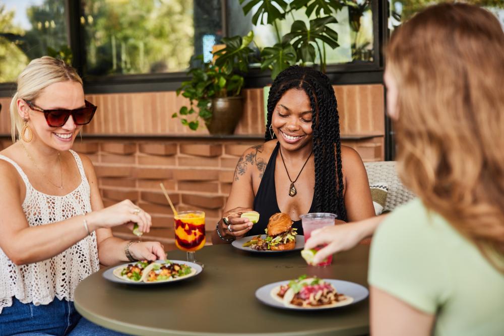 Three women sitting around a table on an outdoor patio with plates of tacos and colorful drinks.