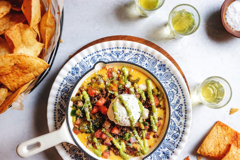 Skillet of loaded queso topped with ground beef, pico de gallo, sour cream and guacamole. The queso is sitting on an ornate plate surrounded by shots of tequila and homemade tortilla chips.