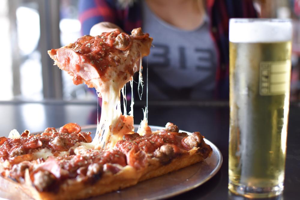 Photo of a person holding a slice of pepperoni and sausage pizza towards the camera. A pale lager beer is on the table