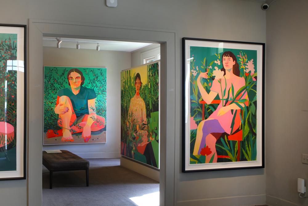 Image of the inside of an art gallery with colorful portraits hanging on the wall, each featuring a woman sitting among greenery and flowers.
