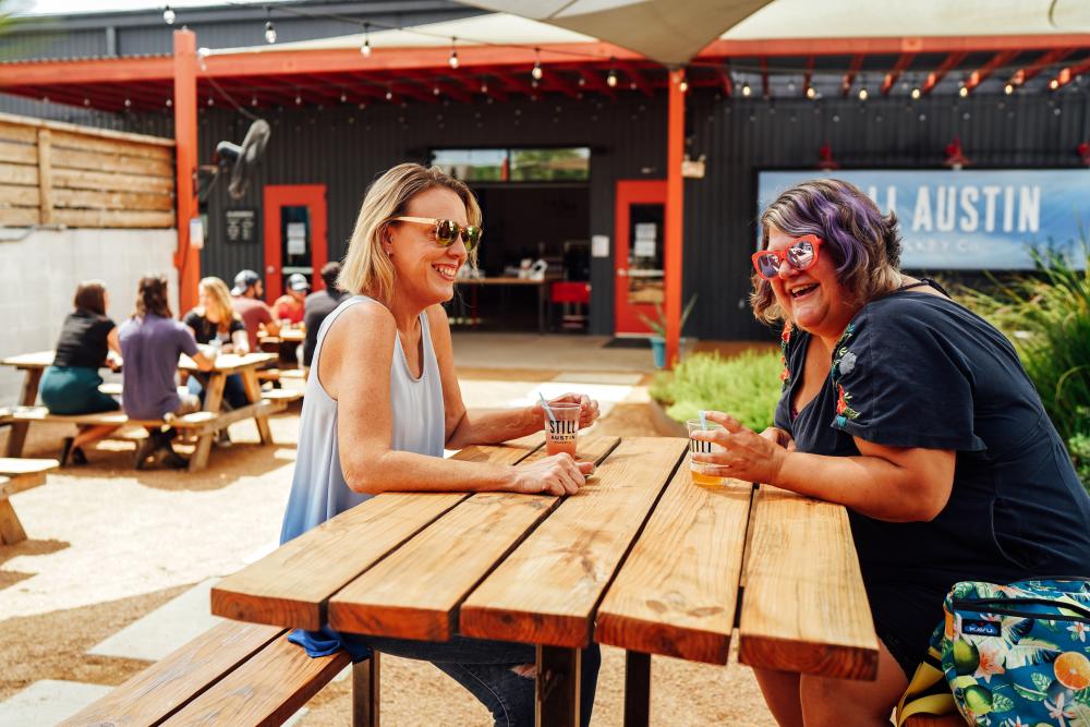 Two women sitting at a picnic table in the Whiskey Garden at Still Austin with cocktails in hand.