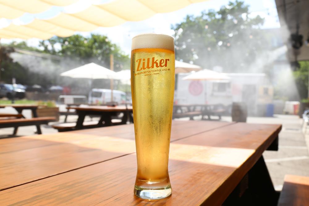 Photo of a tall glass of pale beer on a patio table at Zilker Brewing Company. There is a sun shade above and misting fans in the background