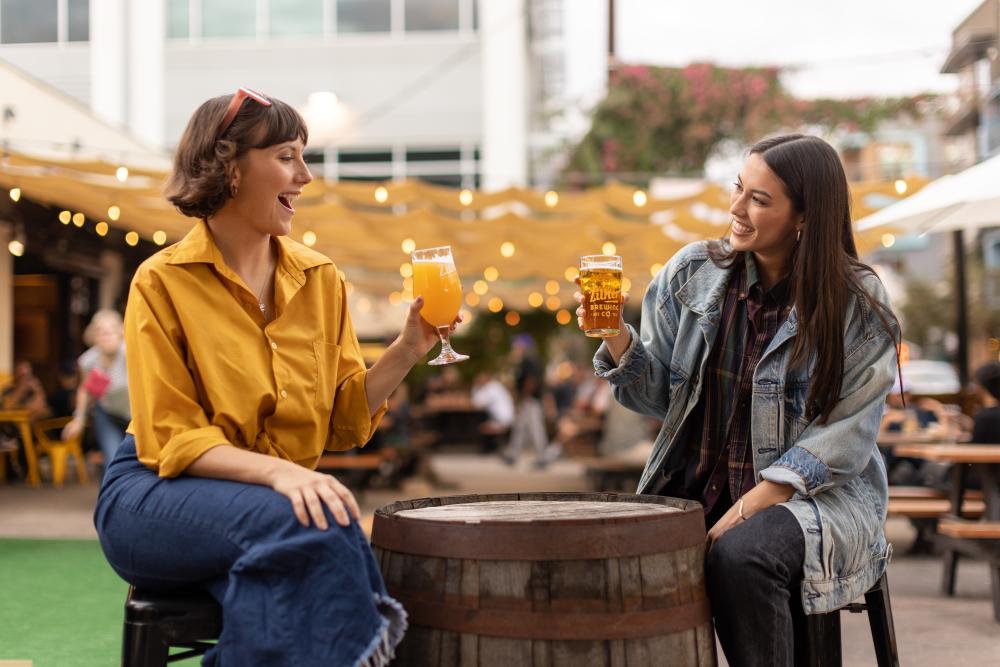 Two women sitting at a bartop at Zilker Brewing Company cheers with glasses of beer