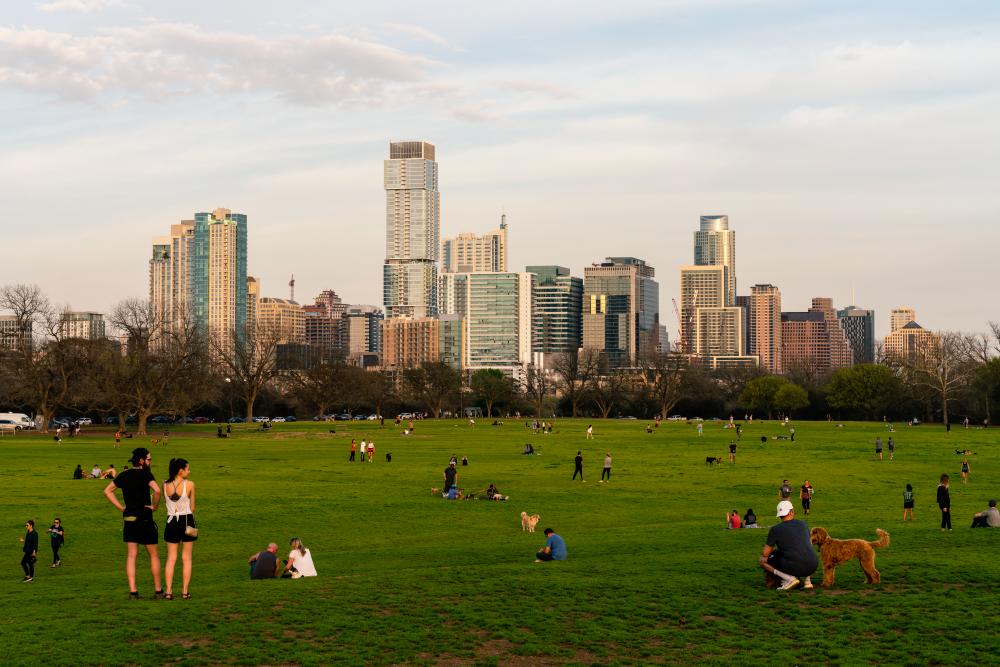 People and dogs are spaced out at the large grassy space at Zilker Park. In the background, the downtown Austin skyline is visible in late afternoon light
