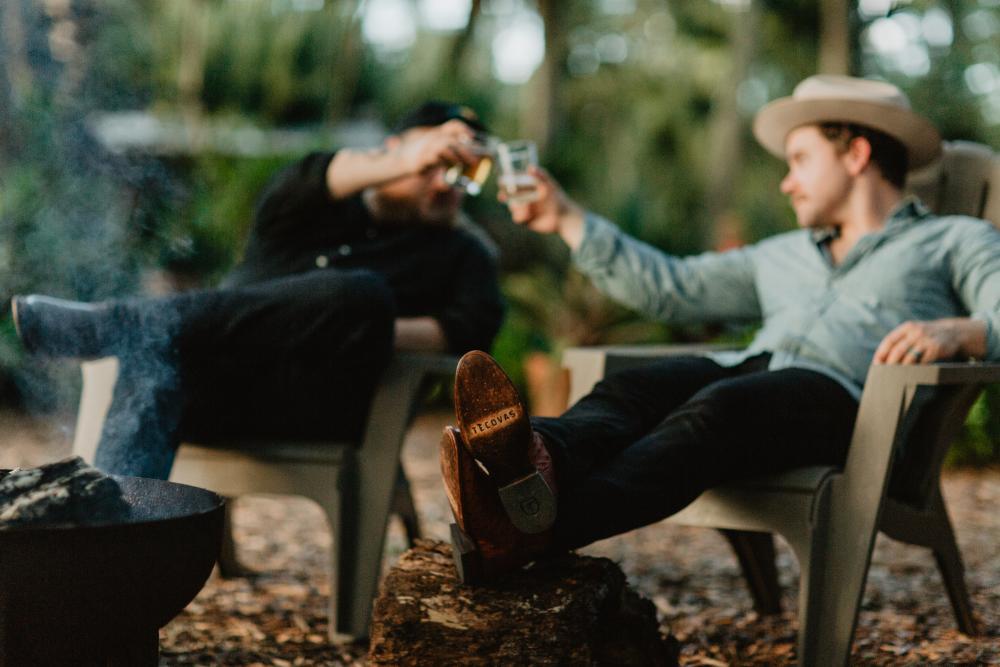 Two men in cowboy boots cheersing whiskey glasses over a fire.