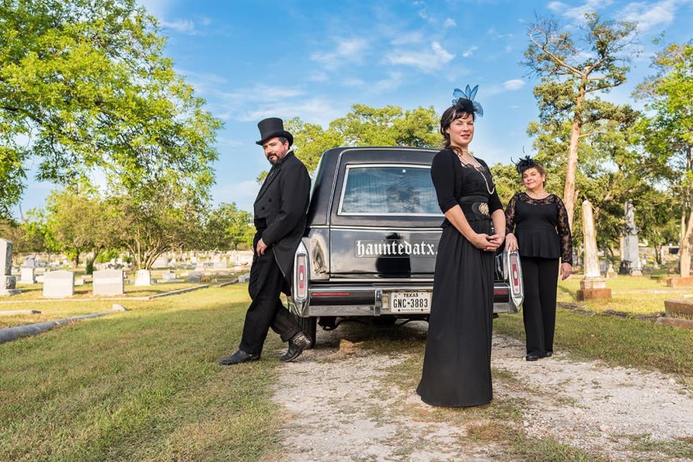 Photo of two women and a man standing in a cemetery and leaning up against a black hearse which says Haunted ATX