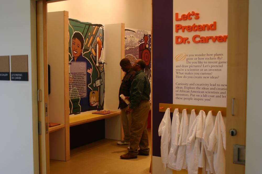 Two men look at an exhibit inside the George Washington Carver Museum. A wall in the foreground has text reading Let's Pretend Dr. Carver... with white lab coats hanging on a rack