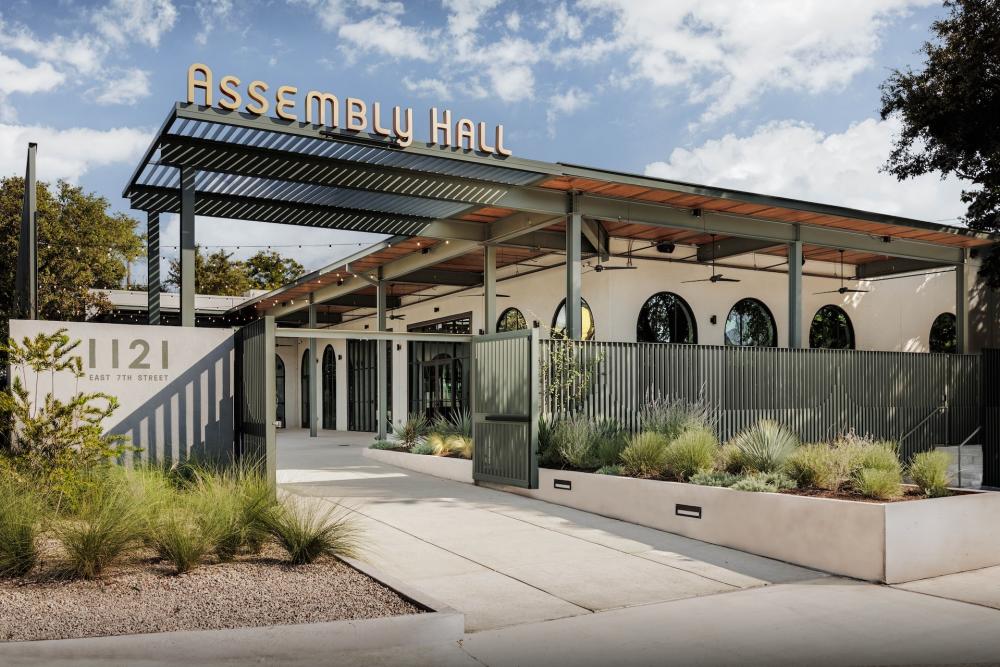 Exterior of an event venue with sign on the roof reading Assembly Hall. The building has a modern facade with dark green fencing, a large patio and native Texas landscaping