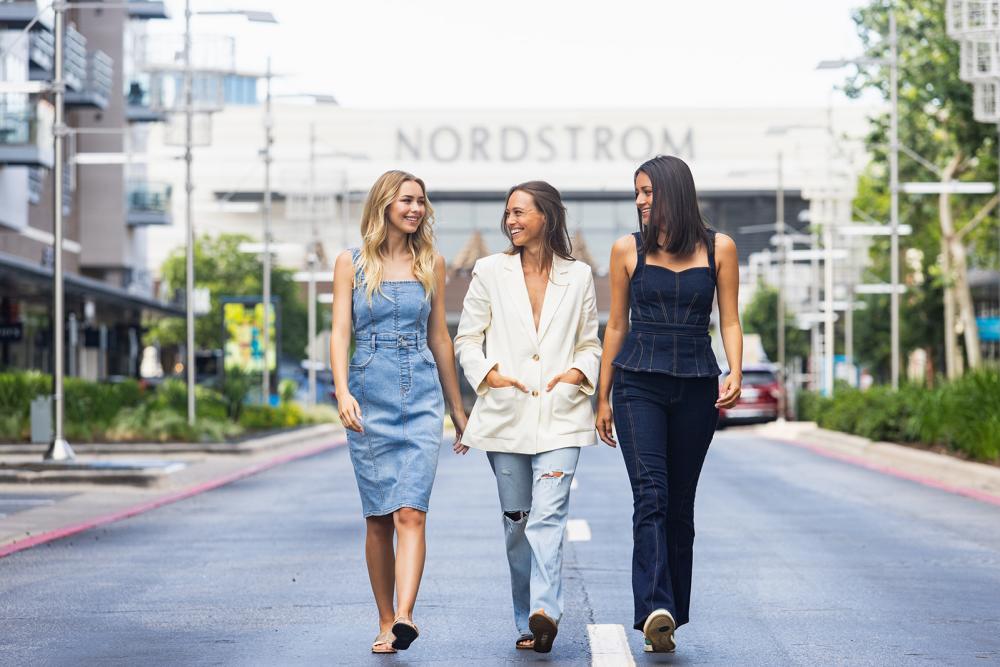 Three women in dressy-casual outfits walking shoulder to shoulder on a street in front of a Nordstrom.