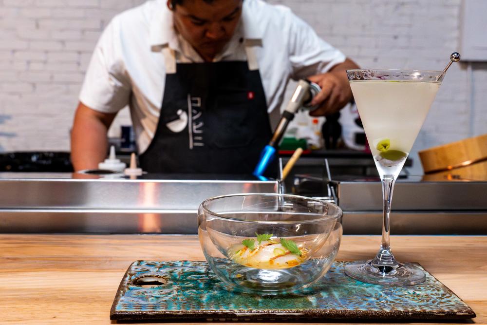 A small bowl of a prepared sushi dish and a cocktail sitting on the bar top with a man in an apron standing in the background cooking.