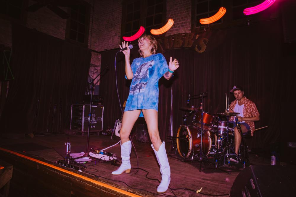 Woman wearing an oversized t-shirt as a dress with tall, white cowboyboots singing on the wooden stage at Stubb's.