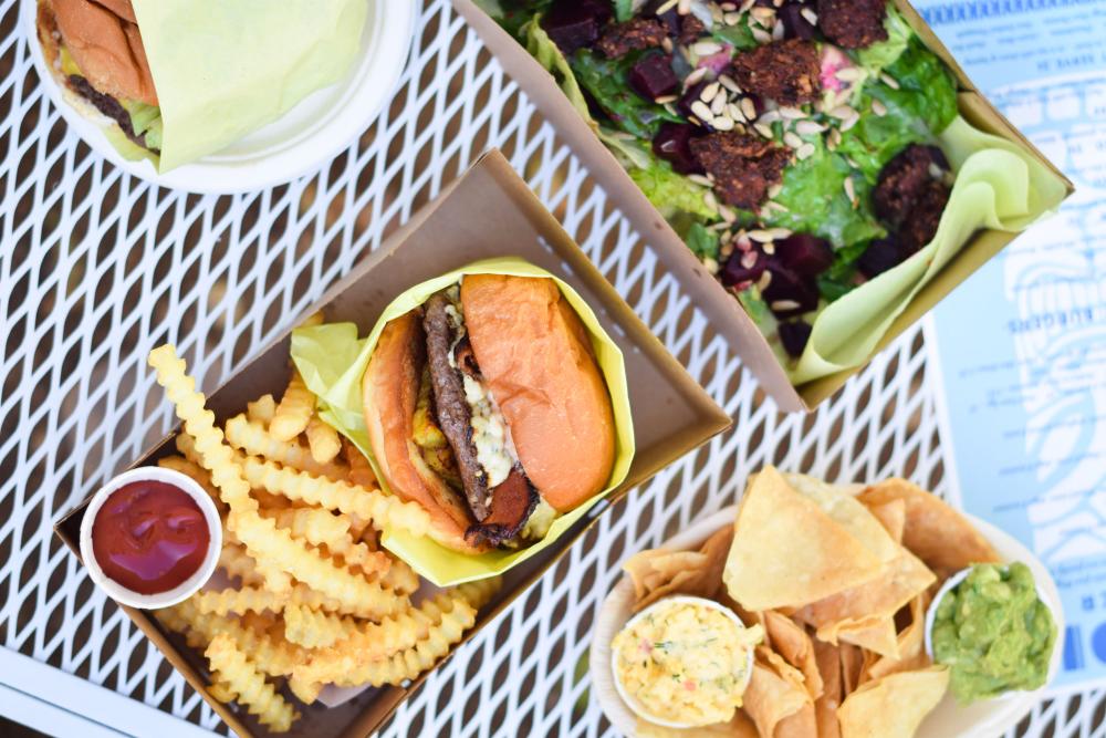 Smash burger with fries, chips and guacamole and beet salad in cardboard containers on a metal grate picnic table.