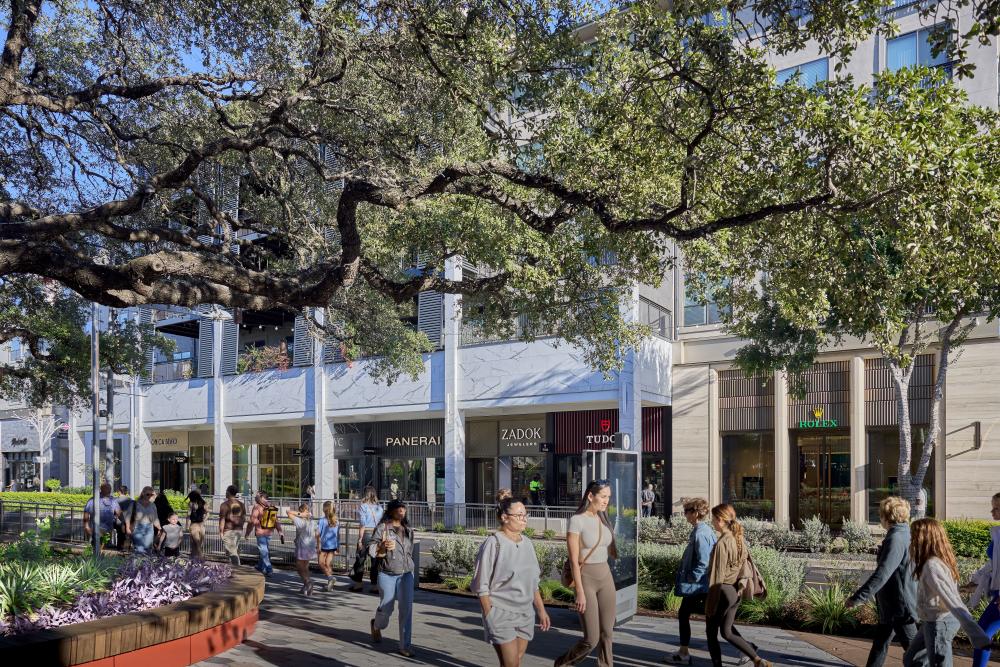 People walking on a sidewalk at The Domain shaded by large oak trees with stores in the background.