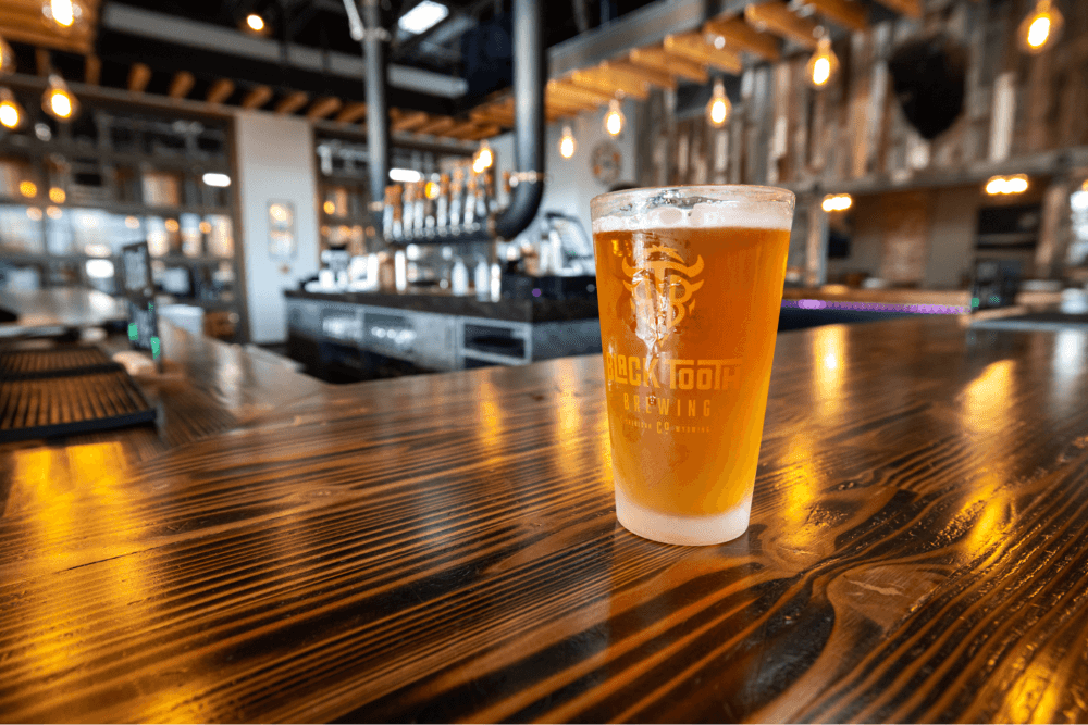 A pint of hazy beer with the Black Tooth Brewing Company logo prominently displayed, resting on a polished wooden bar with the brewery's interior in the background.