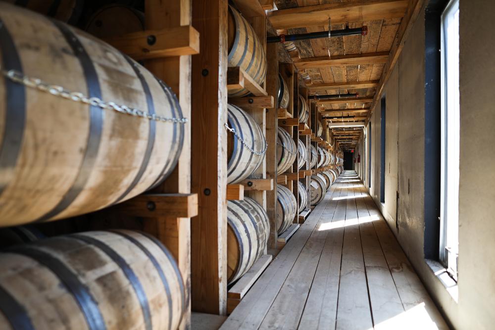 Rows of bourbon barrels line the left side of the photo in a wooden rickhouse.