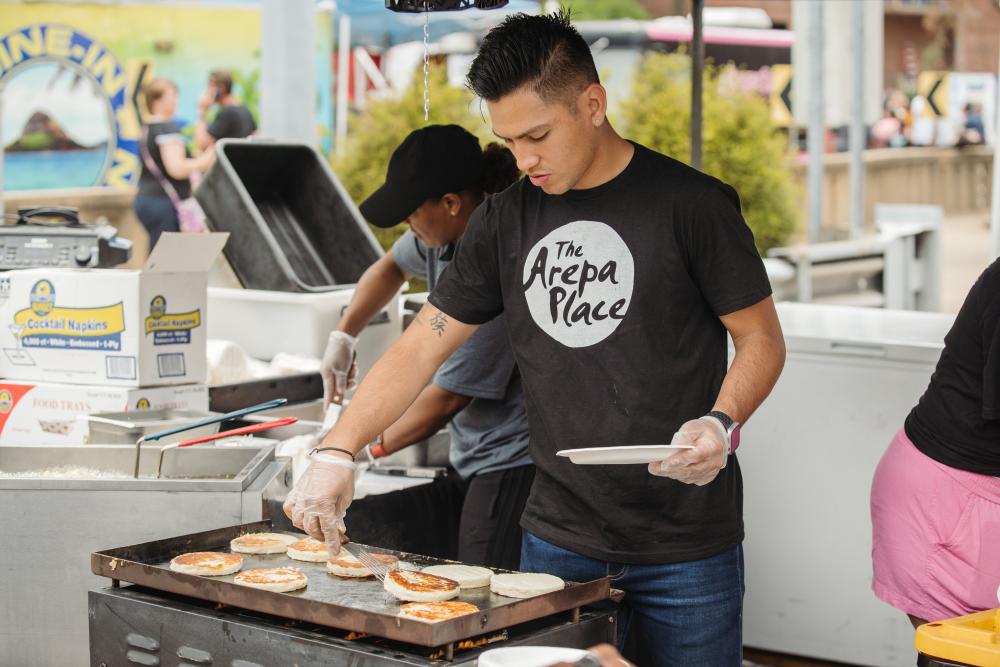 A person wearing a black shirt that says 'The Arepa Place" tends to food on a griddle at Taste of Cincinnati.