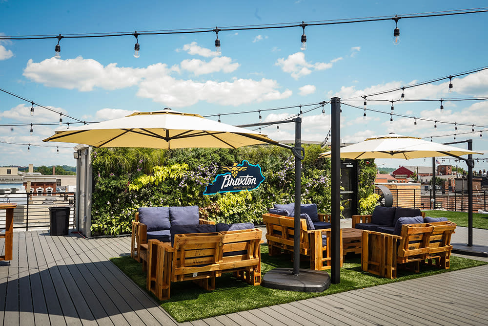 A patio set, umbrellas and string lights sits on a rooftop deck in front of a living plant wall that says Braxton