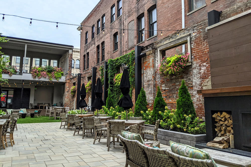 Patio tables are surrounded by a fireplace, greenery and string lights, as well as the brick side of a next door building