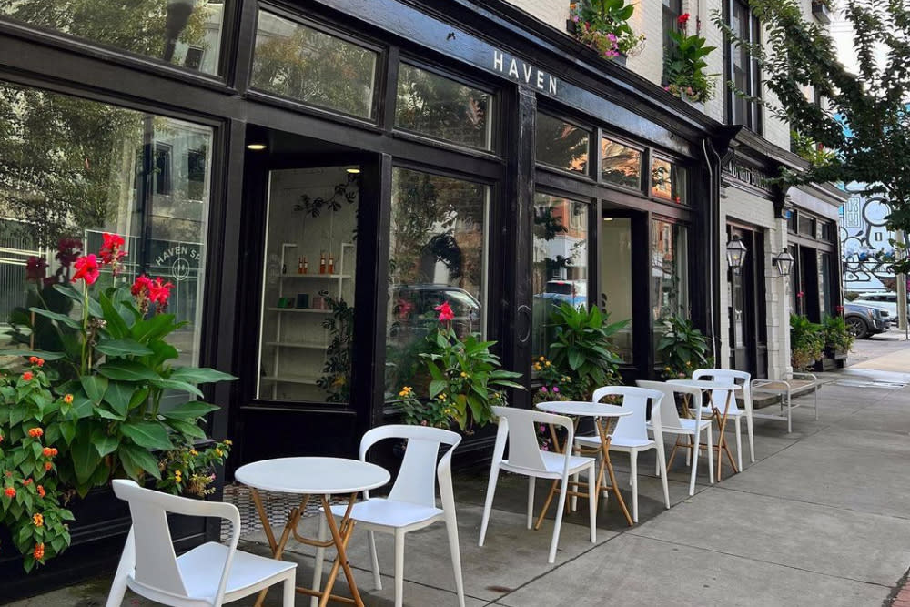 White patio chairs and white patio tables sit on a sidewalk outside a coffeshop called Haven Cafe, with pink flowers