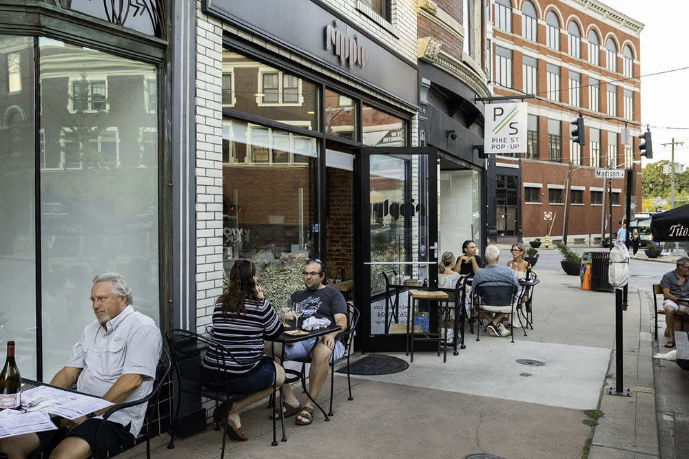 A group of people sits at black metal patio tables on a sidewalk outside Ripple Wine Bar