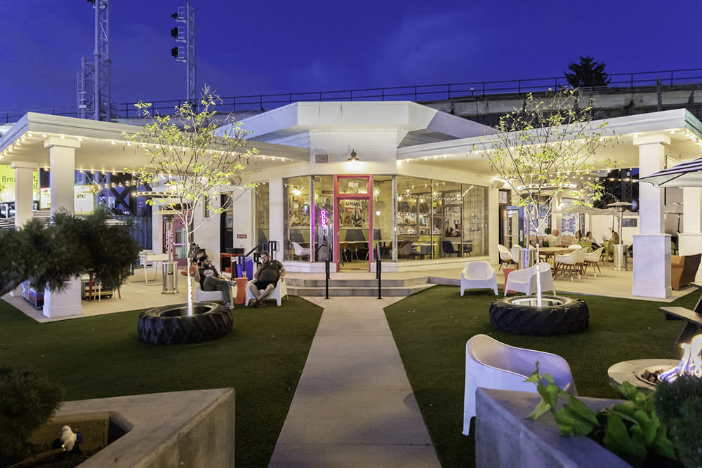 A nighttime view of a former gas station turned bar, with an astroturf yard, pink door and white chairs