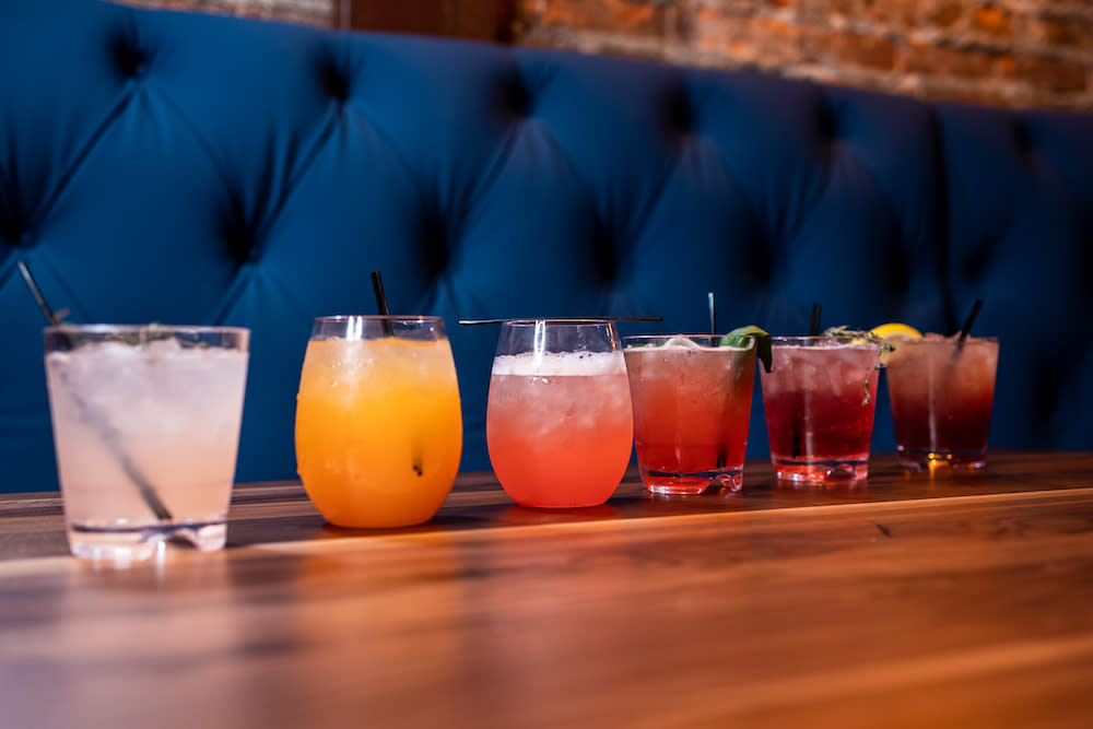 Image is of 6 different cocktails lined up in a row on a wood table with a blue velvet booth seat in the back ground.