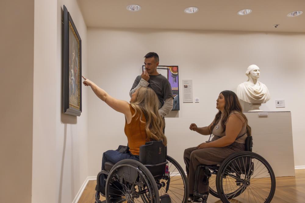 Two women in wheelchairs and a tall man admire and point at a painting in the Kalamazoo Institute of Arts.