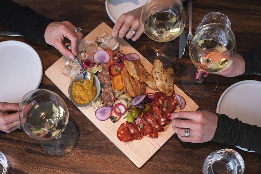 Three sets of hands reach for food from a charcuterie board filled with meats, vegetables, and fruit at the Modest Butcher