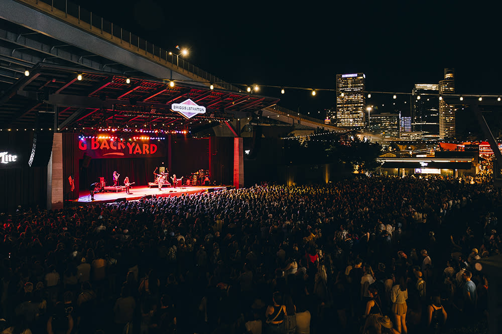 A large crowd gathers at night under a highway overpass for a live outdoor concert at the Briggs & Stratton Big Backyard stage at Summerfest in Milwaukee. The stage is lit with vibrant red and yellow lights as a band performs. String lights hang above the crowd, and the downtown Milwaukee skyline glows in the background.