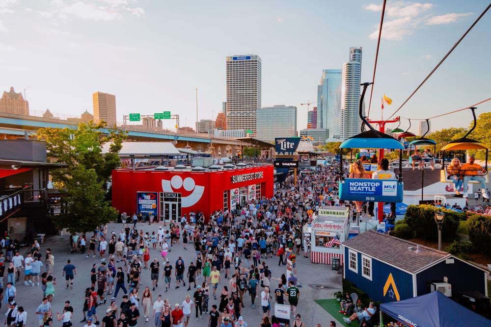 A large crowd moves through the bustling Summerfest grounds in Milwaukee during the day, with the downtown skyline visible in the background. A red building with the Summerfest smiley-face logo serves as the festival store. Above, colorful gondolas on the Skyglider transport people across the venue. The scene is filled with booths, food stands, and signage under a clear sky.