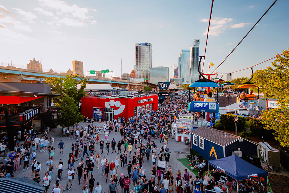 A large crowd moves through the bustling Summerfest grounds in Milwaukee during the day, with the downtown skyline visible in the background. A red building with the Summerfest smiley-face logo serves as the festival store. Above, colorful gondolas on the Skyglider transport people across the venue. The scene is filled with booths, food stands, and signage under a clear sky.