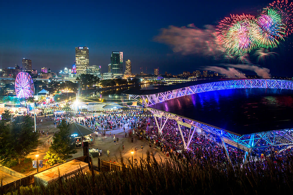 A vibrant nighttime scene at Summerfest grounds in Milwaukee, featuring a large crowd, illuminated Ferris wheel, and the glowing roof of the American Family Insurance Amphitheater. The downtown skyline sparkles in the background as colorful fireworks burst over Lake Michigan, adding to the festive atmosphere.