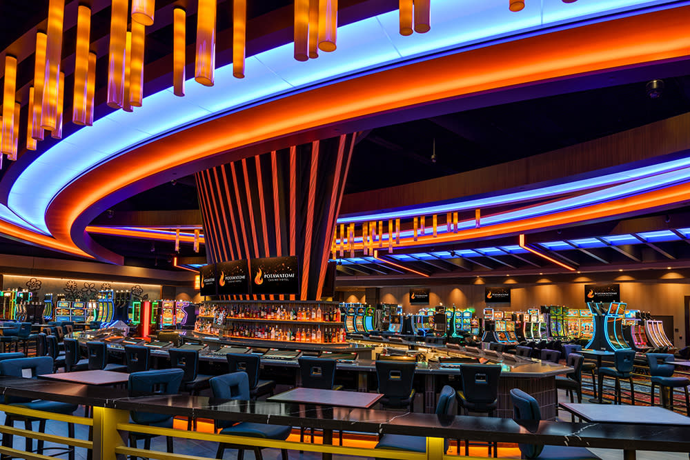 A vibrant, modern casino interior at Potawatomi Casino | Hotel in Milwaukee. The image features a sleek central bar beneath a dramatic, circular ceiling structure with glowing orange and blue LED lighting. Around the bar, rows of gaming tables and slot machines extend across the floor. Hanging orange cylindrical lights add to the atmosphere, and plush blue chairs line the tables and slots. The space feels energetic and upscale.