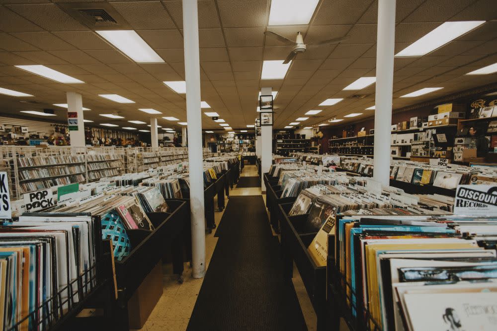 A wide view inside a record store shows long rows of vinyl bins stretching toward the back, organized by genre and artist. Fluorescent lights line the ceiling above white support columns, while shelves along the walls display more records and audio equipment.