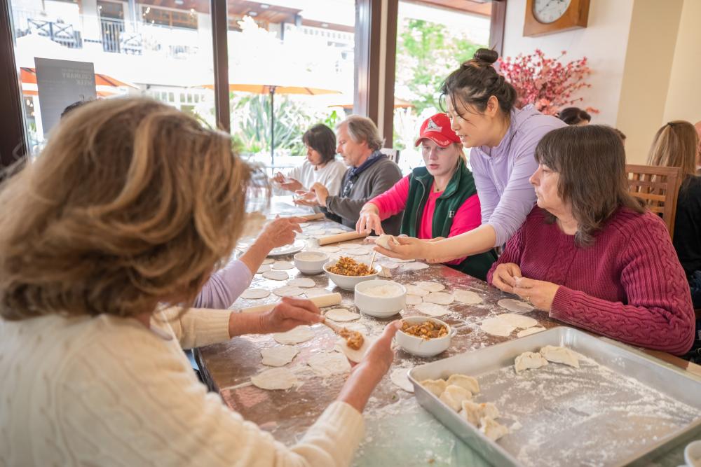 This is an image of a group of people sitting around a flour-ladden table making potstickers. There is one woman standing that is teaching the group how to make the potstickers during Carmel Culinary Week in Carmel-by-the-Sea